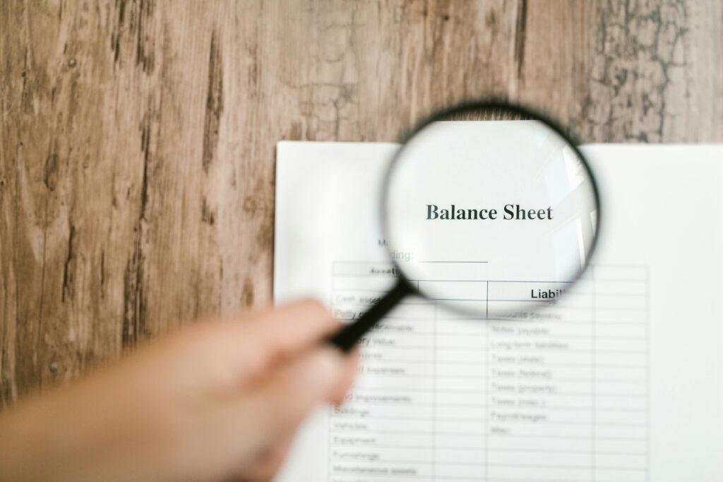 A hand holds a magnifying glass over a balance sheet, which is laying on a wooden table.