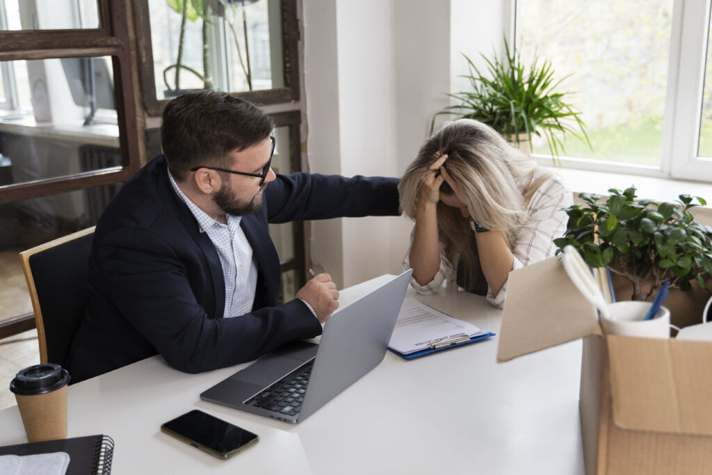 A woman sits at a desk with her head in her hands to signal she’s gotten bad news, while a man tries to comfort her.