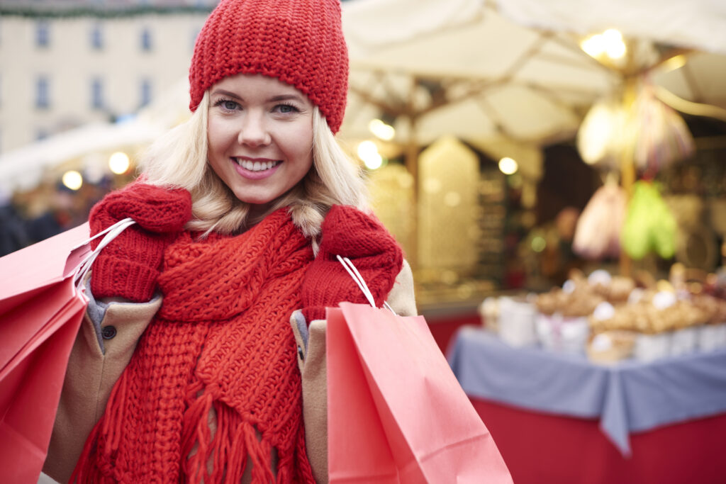 A woman in a red toque and mittens holds two shopping bags, standing in front of some kind of business with fairy lights.