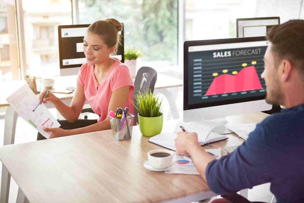 Man and woman sit at a desk to analyze charts, with one computer screen saying “Sales Forecast”.