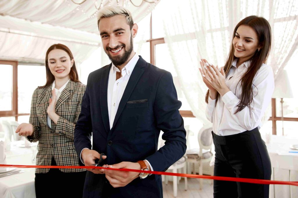 Man cuts red ribbon for a grand opening of a business, with two women applauding behind him