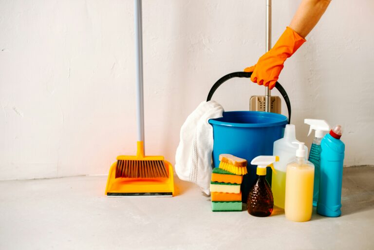 A person wearing an orange rubber glove holding a blue bucket filled with cleaning supplies next to a yellow broom and dustpan.